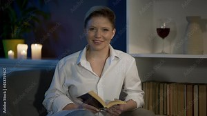 Comfort concept. Pleasant pastime in evening. Rest from work. Woman smiling looking into camera. Oficce worker reads book by candlelight in cozy apartment. High quality 4k footage.