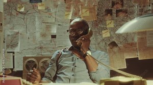 Experienced Black detective sitting at desk with investigation map behind and talking during call on old corded telephone in the office at night