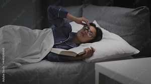 Tired middle aged woman falling asleep while reading book in bed. Caucasiana brunette in eyeglasses ad pajamas lying in bed with soft pillows and blanket.