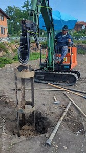 A man on a mini excavator drilling holes for piles at a construction site. The excavator's boom and auger are in action, while the operator controls the process with the levers.