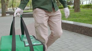 A male delivery man wearing white medical gloves delivers orders safely in a thermocouple down the street on foot. A close-up of the courier’s hand and delivery bag.