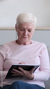 Happy senior Caucasian woman daydreaming and making notes, creating shopping list or planning agenda in notebook at home. Vertical video.