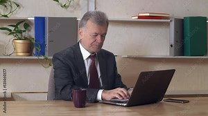 Old man in years surfing internet and typing on keyboard laptop in contemporary office. Old-aged boss working with computer. Grey headed manager wearing in black fashionable suit reading document