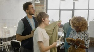 Professional dressmaker showing fabric and telling about its advantages to female client choosing material for a dress in atelier, African-American assistant taking notes during conversation
