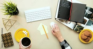 Top view on Cauasian female hands typing on keyboard and tapping with mouse while working at computer. View from above on desktop of businesswoman drinking hot coffee with cookies.