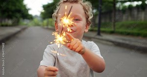 Young child holds handheld firework, pointing excitedly at two sparkling flames. Mesmerized by magic in his hands, his face radiates innocence and wonder. Toddler expression reflects pure delight