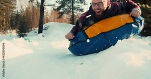 WINTER SNOW FUN. Excited happy 20s man enjoying sledging down slope on inflatable sled ride in sunny forest slow motion.