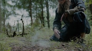 Concentrated elf with pattern on face in mystical costume makes smoke offering to forest ghosts sitting on grass in ancient wood closeup slow motion