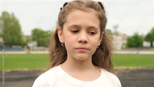 Sad pensive beautiful young girl with long hair looking at the camera with a thoughtful expression. Beautiful preteen female standing alone outdoors in a park with a serious face