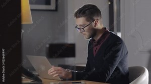 Multitasking. Handsome young man wearing glasses and working withlaptop while sitting in modern office. Freelancer in coworking at night Stock Video