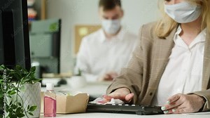 Employee woman wearing medical face mask wiping keyboard with antibacterial cloth, using hand antiseptic before typing, working at computer. Office work during the covid epidemic. Safety protection.