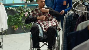 Middle eastern man in wheelchair choosing clothes to try on in the fitting room, asian female employee at the store helping him and providing a great shopping experience. Camera A.
