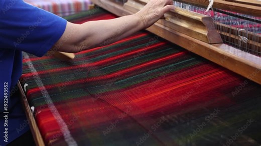 Close-up of a craftsperson weaving vibrant red and green tartan fabric on a traditional wooden loom. Hands guide the shuttle and threads in a textile workshop setting