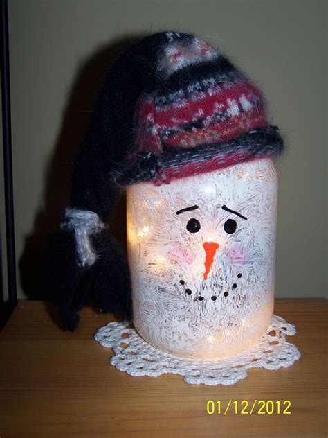 A Lit Up Snowman Jar With A Hat On Its Head Sitting On A Doily