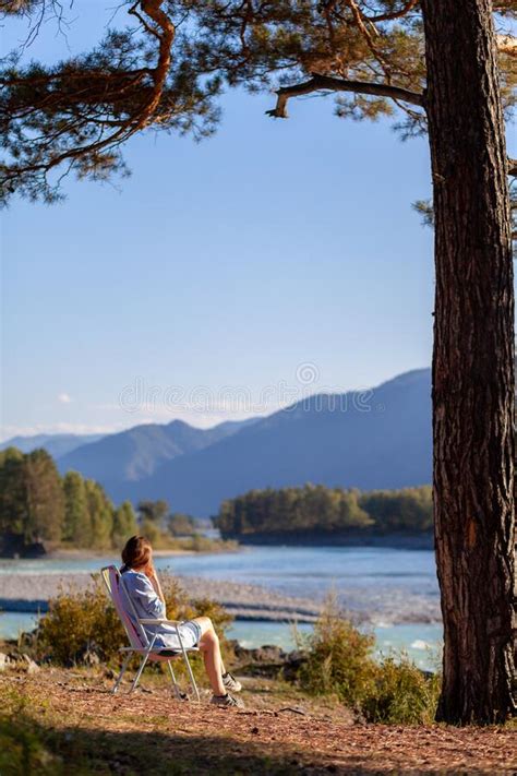 A Woman Is Sitting On A Folding Chair On The Bank Of A Mountain River