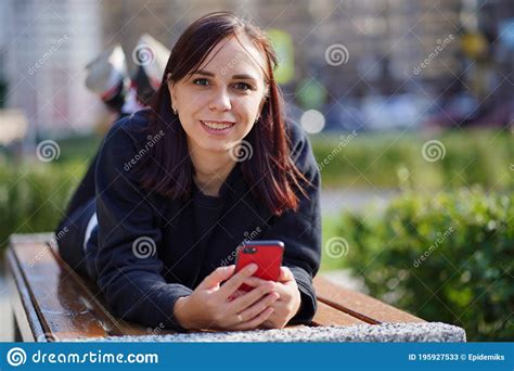 Young Woman Lying On Her Stomach Outside On A Bench With Her Legs Up And Browsing Her Cell