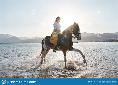 Caucasian Blonde Girl Riding A Horse Through The Water At Sunrise Stock
