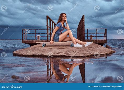 Sensual Girl Sitting On A Skateboard On The Beach Is Enjoying Amazing Dark Cloudy Weather During