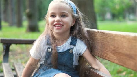 Small Happy Child Girl Sitting On A Bench Resting In Summer Park Stock
