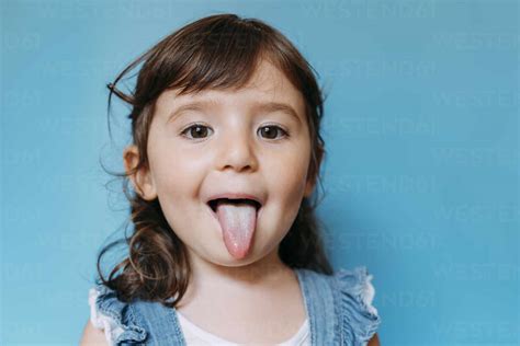 Portrait Of Cute Little Girl With Tongue Out On Blue Background Stock Photo