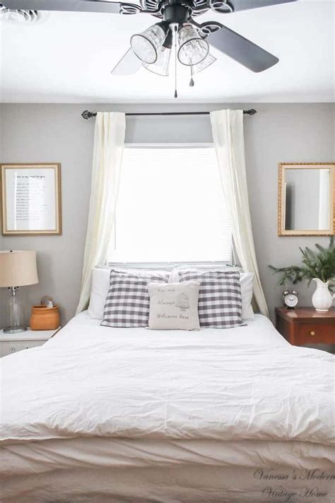 A White Bed Sitting Under A Ceiling Fan In A Bedroom Next To A Window With Two Framed Pictures