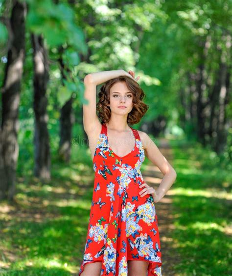 Young Brunette Girl In Red Dress Posing On Alley In Summer Park Against
