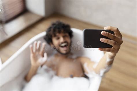 Joyful Indian Man Lying In Bathtub And Waving Hand On Smartphone Webcamera Resting In Bath