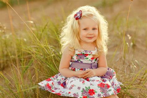Little Curly Blonde Girl Sits And Smiles On Sand And Grass In Sunny