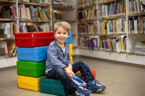 Adorable Little Boy Sitting In Library Reading Book And Choosing What