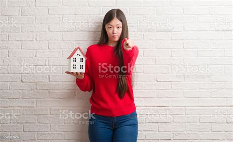 Young Chinese Real State Agent Woman Over Brick Wall Holding House Pointing With Finger To The