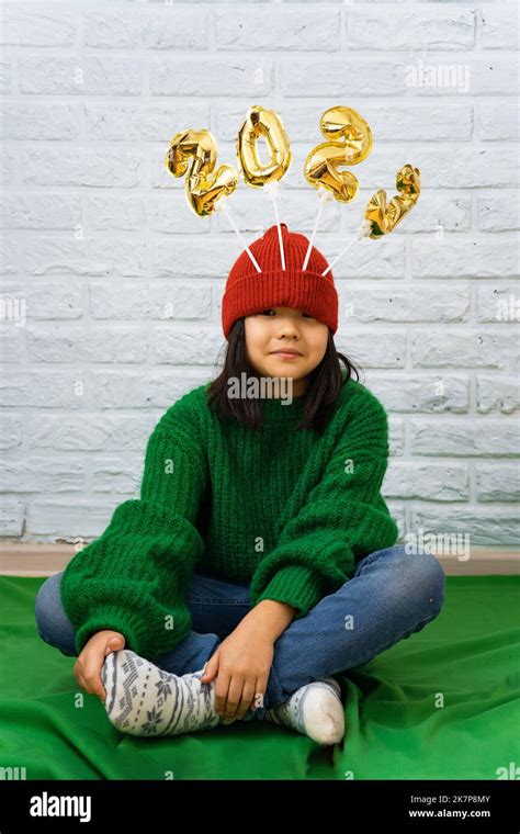 Funny Asian Girl In A Green Sweater Sits On The Floor With Golden Balloons With The Number 2023