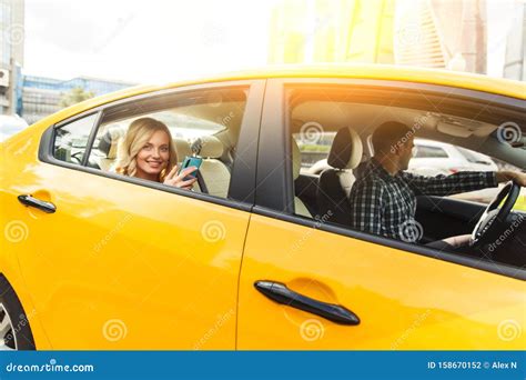 Photo Of Young Blonde With Phone In Her Hand Sitting In Back Seat Of Yellow Taxi With Driver