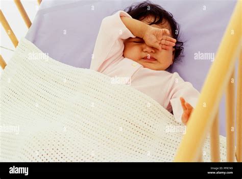Baby Lying On Back In Cot With One Arm Raised And Rubbing Her Eyes Stock Photo Alamy