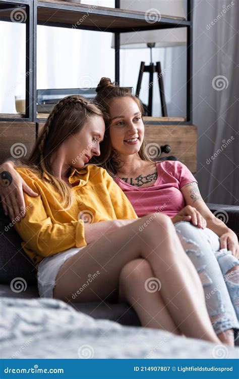 Two Pretty Lesbians Embracing While Sitting On Sofa In Living Room