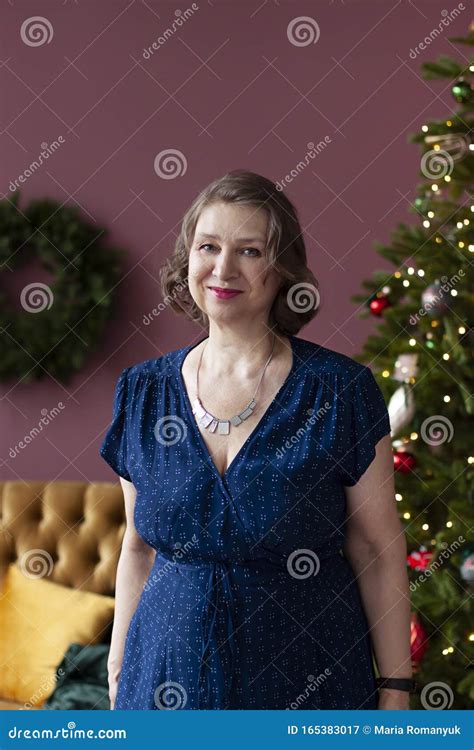 Mature Woman Is Standing And Smiling In The Interior With Christmas Or New Year Festive
