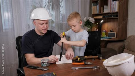A Young Father Teaches His Son To Work With A Saw The Boy Holds The