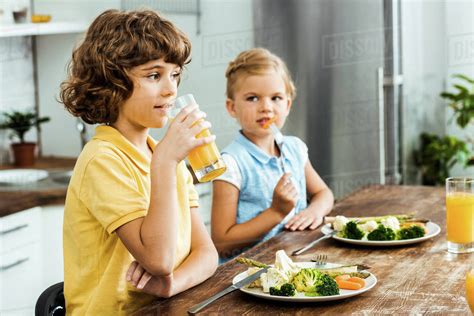 Adorable Kids Sitting Together At Table Eating Vegetables And Drinking
