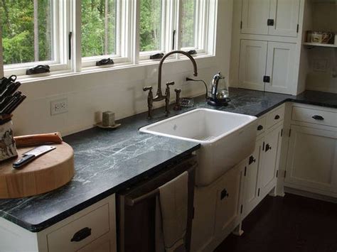 The kitchen with light grey island with dining seating features a black countertop and full kitchen sink and white cabinet. white farmhouse sink with slate counter top, light wood ...