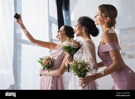 african american bride with bridesmaids holding wedding bouquets while taking selfie on