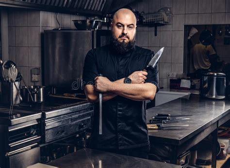 Bearded Cook In Black Uniform Holds Knife And Standing With Crossed Arms In The Kitchen Stock