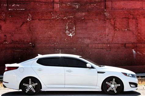A White Car Parked In Front Of A Red Brick Wall With Black Spokes On It