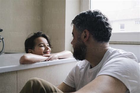 Smiling Daughter Leaning On Bathtub While Looking At Father Sitting In