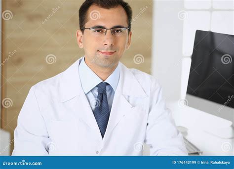 Doctor Man Sitting At The Desk At His Working Place And Smiling At
