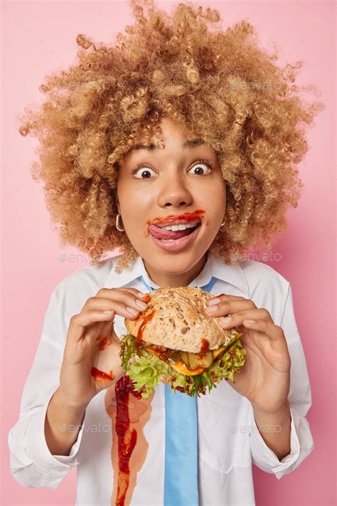 Vertical Shot Of Curly Haired Woman Enjoys Eating Junk Food Licks Lips