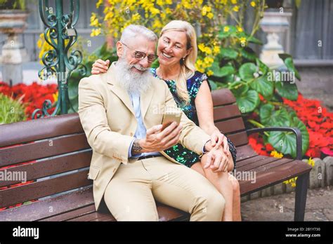 Senior Couple Using Smartphones Outdoor Sitting On A Bench Mature People Having Fun With New
