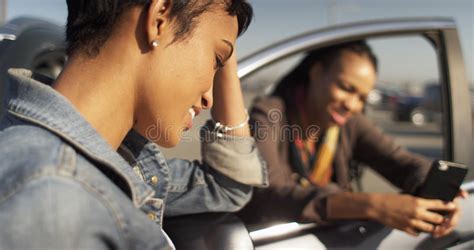 Two Black Women Friends Texting On Cell Phone And Leaning Against Car