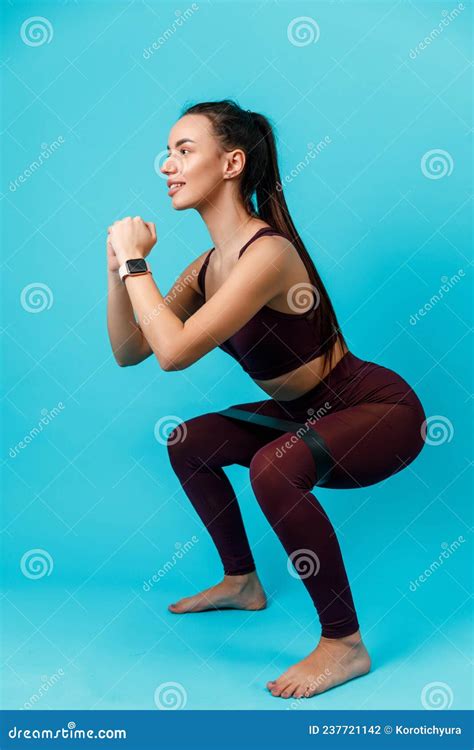 Beautiful Sportive Brunette Woman Squats With Elastic Band On Blue