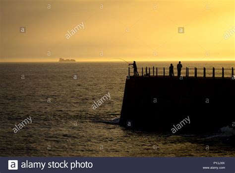 Two People And A Fisherman Stand On A Silhouetted Pier At Sunset Stock