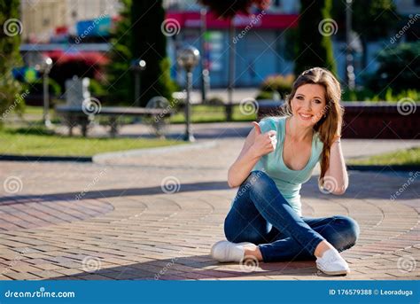 A Young And Pretty Girl In Jeans Sits On A Sidewalk Stock Photo