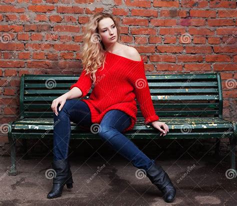 An Incredible Blonde Girl Posing In A Trendy Red Sweater Jeans And Black Boots Sitting On A